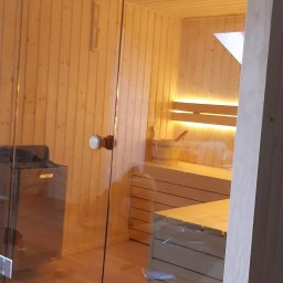 Sauny - Nowoczesna sauna z przeszklonymi drzwiami, drewnianym wykończeniem i podświetlanymi ławkami, widoczny piec elektryczny.