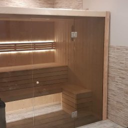 Sauny - Nowoczesna sauna z jasnego drewna z przeszklonymi drzwiami, oświetleniem LED pod ławkami, umieszczona w łazience z beżowymi płytkami imitującymi kamień.