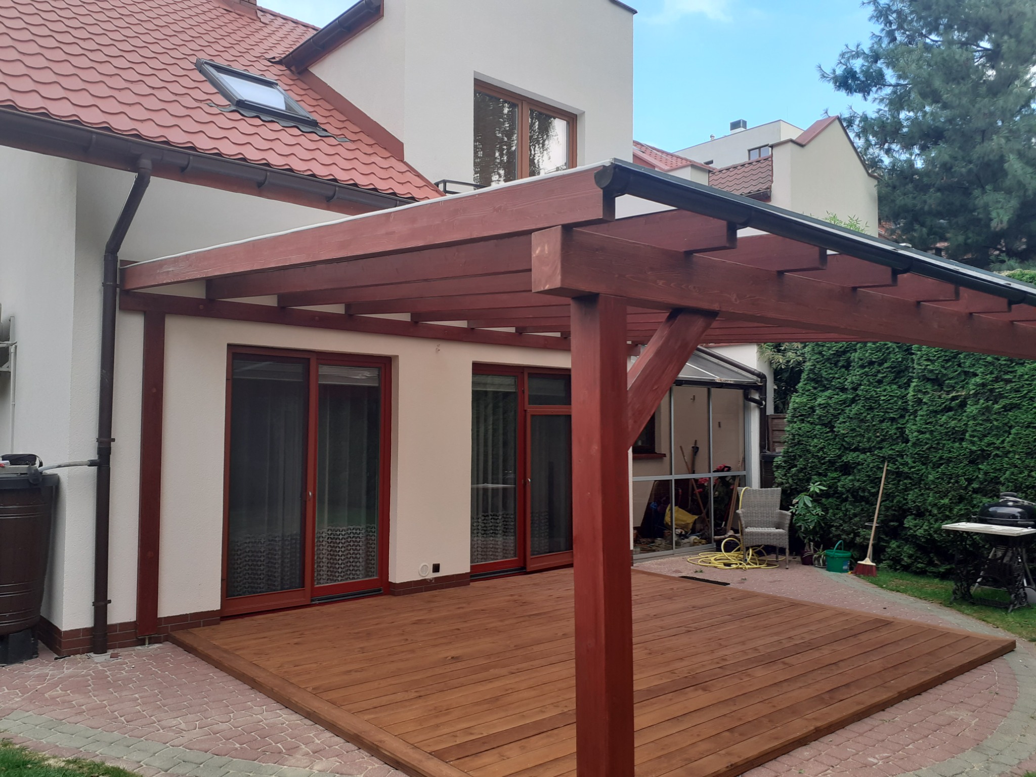 Ciemnobrązowa konstrukcja z belek nad drewnianym tarasem przylegającym do domu z czerwonym dachem i białymi ścianami, widok na ogród z grillem i krzesłem.