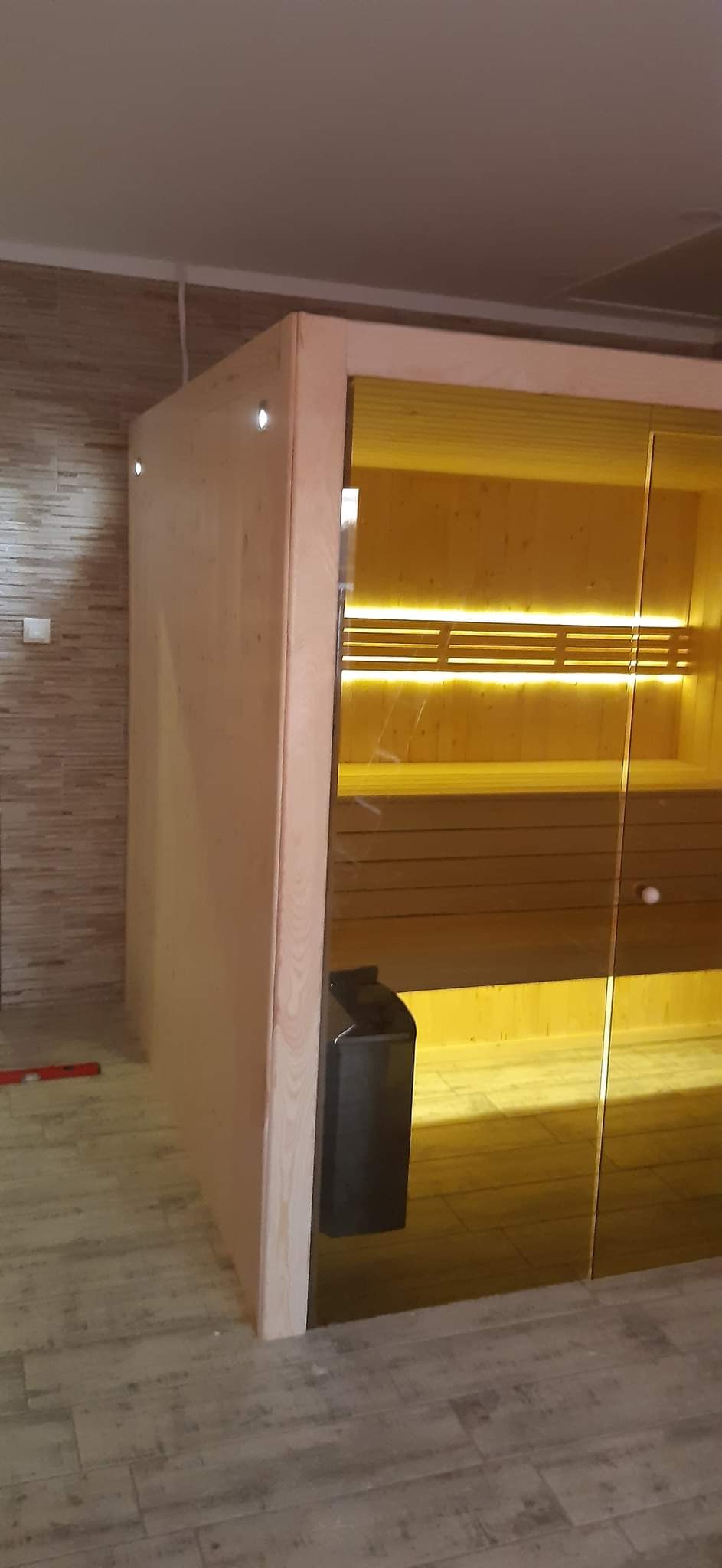 Nowa sauna z drewnianym wykończeniem i przeszklonymi drzwiami, oświetlona od wewnątrz, z widocznym piecem.