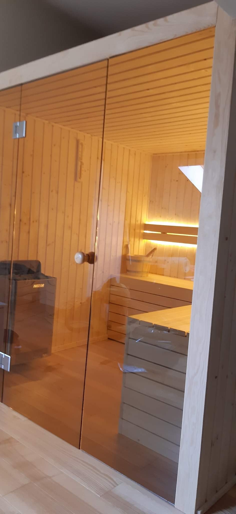 Nowoczesna sauna z przeszklonymi drzwiami, drewnianym wykończeniem i podświetlanymi ławkami, widoczny piec elektryczny.
