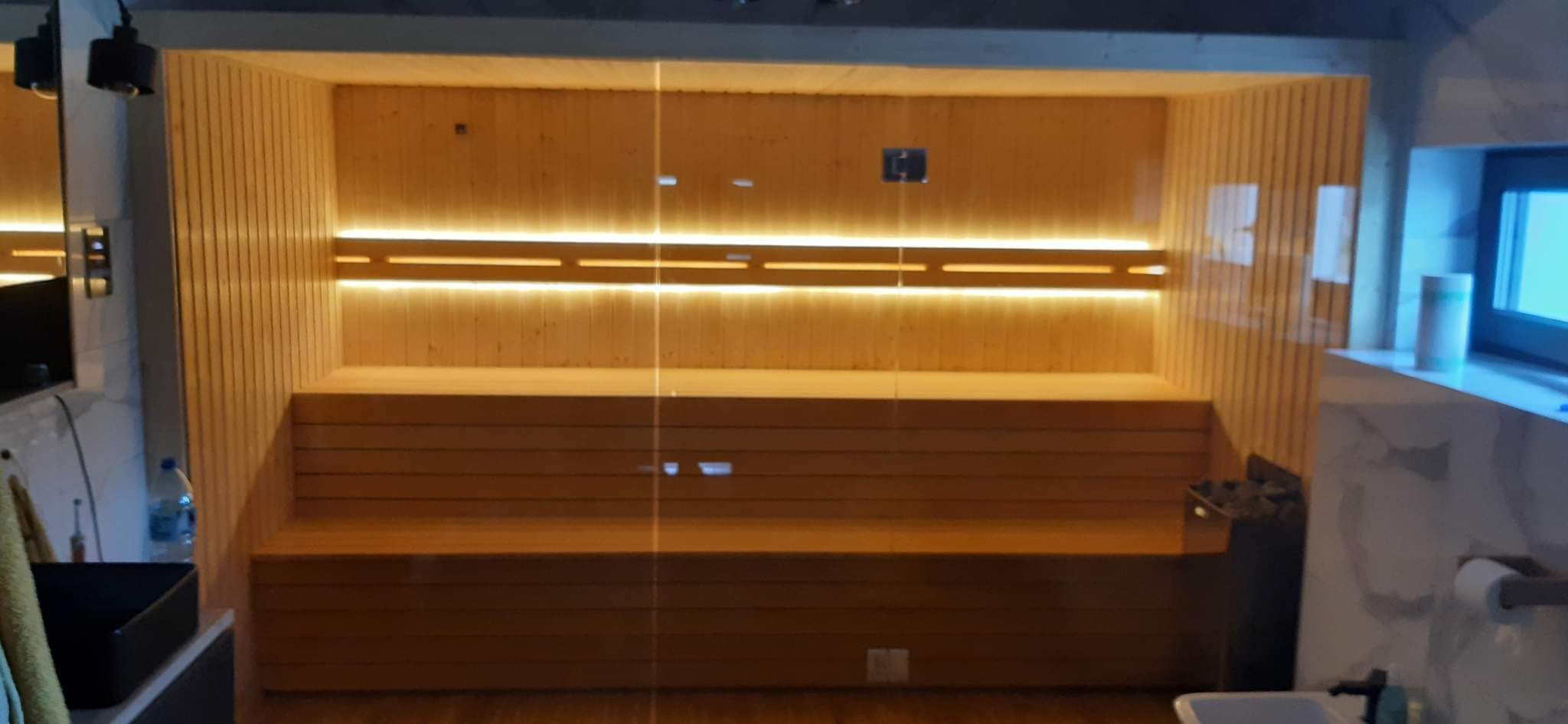 Wnętrze sauny z drewnianymi ławkami, oświetleniem LED i szklanymi drzwiami, widok z zewnątrz sauny na marmurowe wykończenie łazienki z oknem