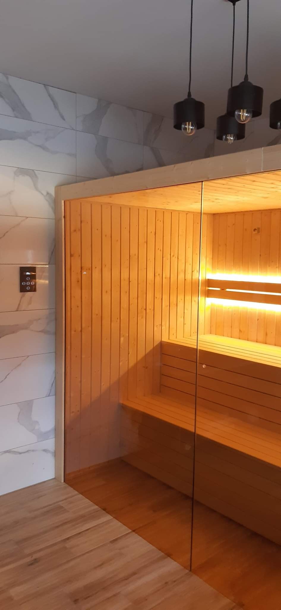 Nowoczesna sauna z drewnianym wykończeniem, przeszklonymi drzwiami i marmurowymi płytkami na ścianie, oświetlona ciepłym światłem.