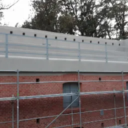 Odrestaurowany ceglany mur fortu z metalowymi okiennicami i nową betonową nadbudową z metalową balustradą, widoczne rusztowania.