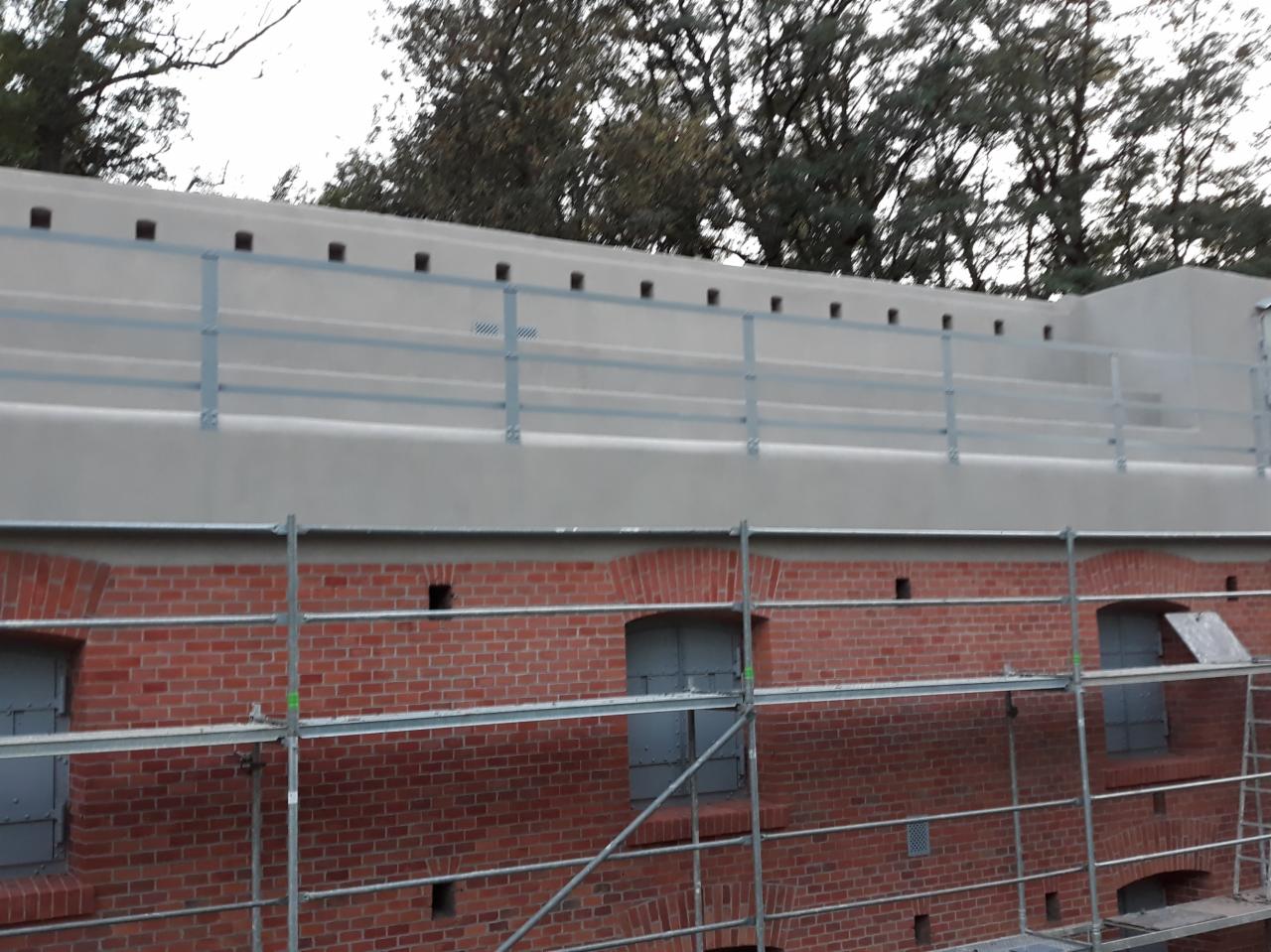 Odrestaurowany ceglany mur fortu z metalowymi okiennicami i nową betonową nadbudową z metalową balustradą, widoczne rusztowania.