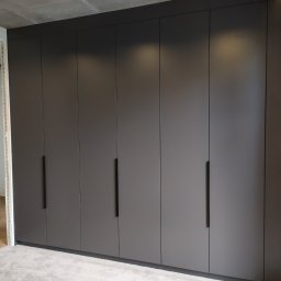 Pracownia Meblarska - Duża, matowa, szara szafa wnękowa z minimalistycznymi, długimi uchwytami, w sypialni z betonowym sufitem i jasną wykładziną.