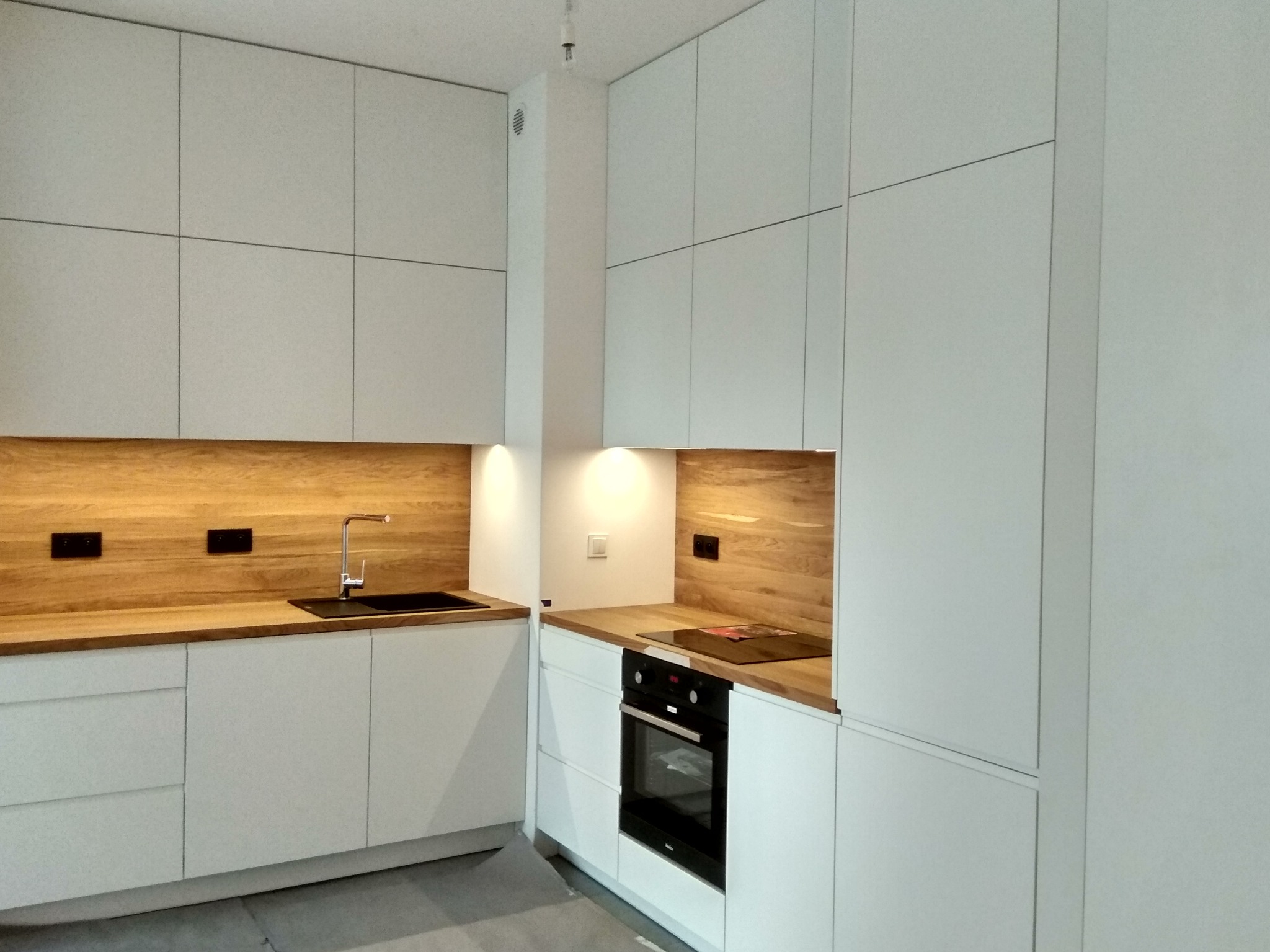 Nowoczesna, minimalistyczna kuchnia z białymi, gładkimi frontami szafek i drewnianymi blatami oraz panelami ściennymi, wyposażona w czarny zlew i płytę indukcyjną.
