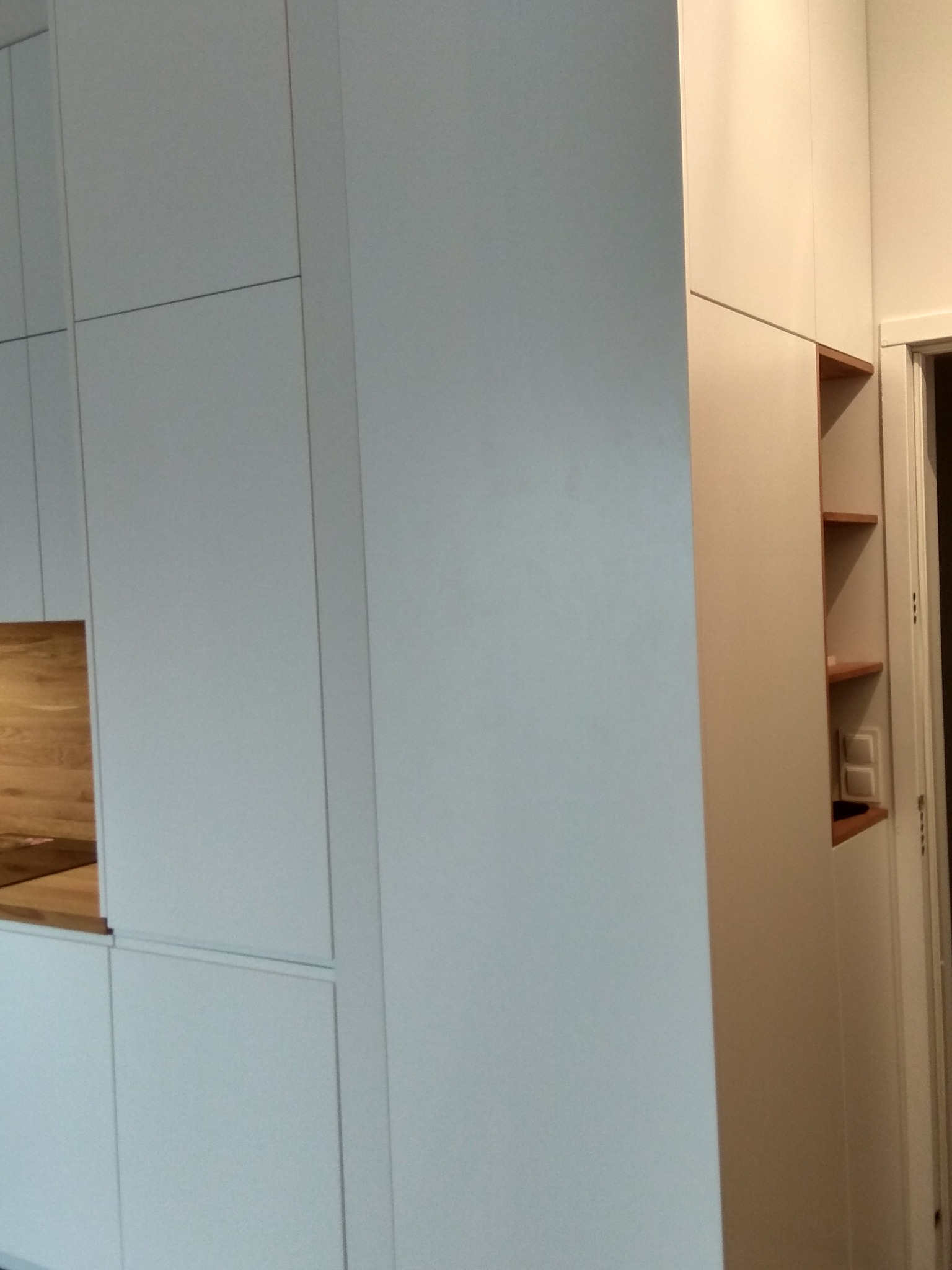 Biała zabudowa meblowa z drewnianymi półkami wnękowymi, widoczne minimalistyczne fronty szafek i fragment blatu.