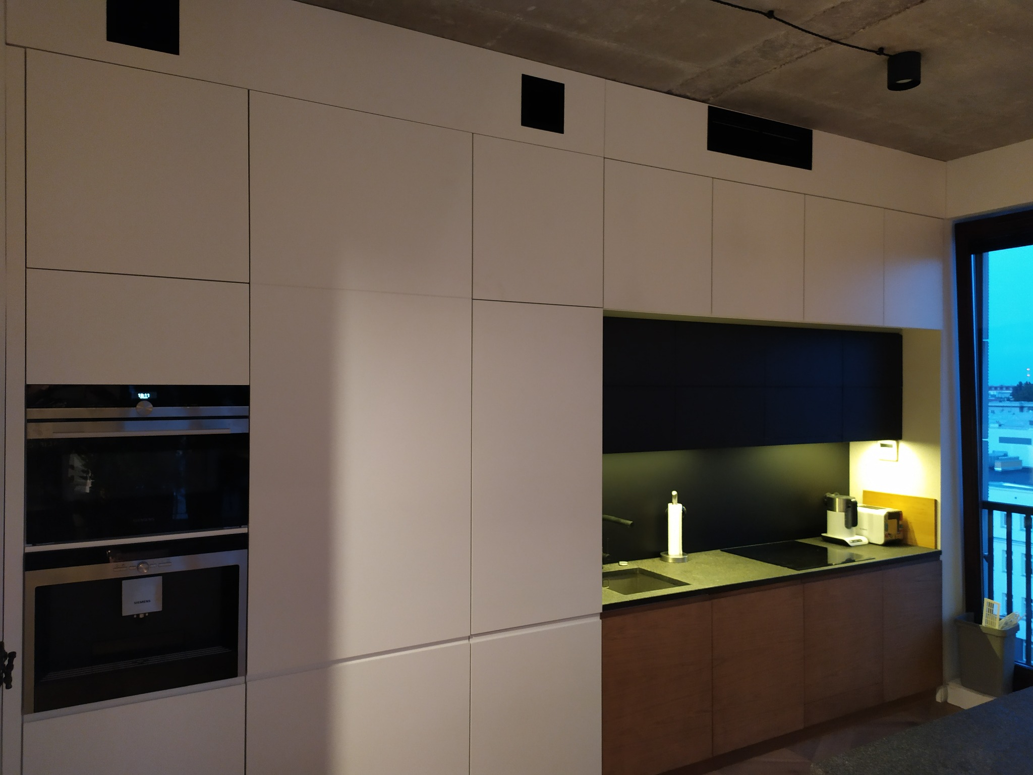 Minimalistyczna kuchnia z białymi frontami szafek, drewnianymi dolnymi szafkami, czarnym blatem i sprzętem AGD marki Siemens, oświetlona punktowo, z widocznym betonowym sufitem.