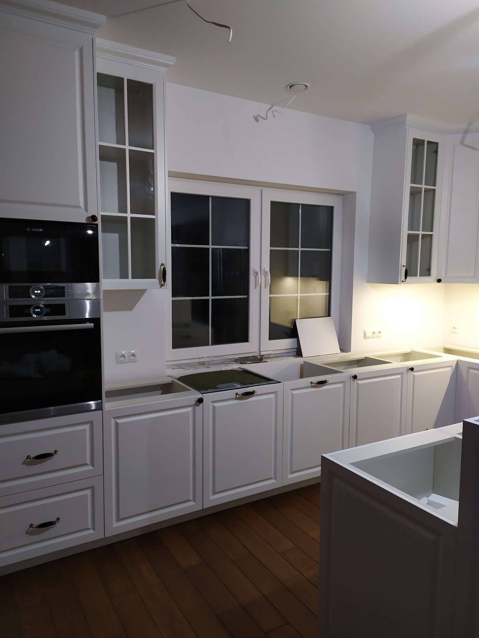 Biała, klasyczna zabudowa kuchenna w trakcie montażu, widoczne szafki dolne i górne z przeszklonymi frontami, brak blatu, oświetlenie podszafkowe, sprzęt AGD marki Bosch, drewniana podłoga, okno...