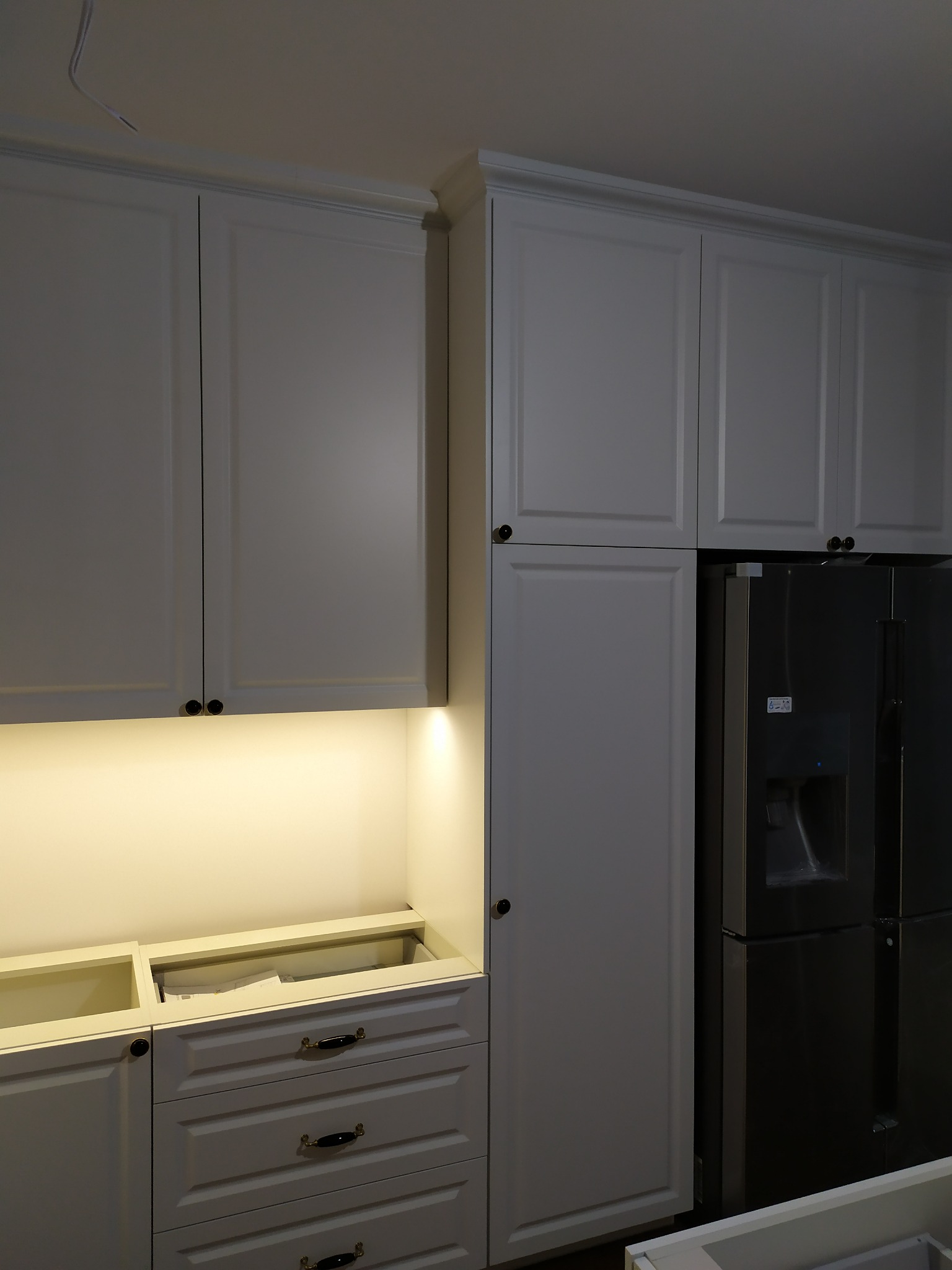 Białe szafki kuchenne z czarnymi uchwytami, podświetlane blaty, widoczna lodówka w zabudowie, brakujące blaty robocze, instalacja elektryczna na suficie.