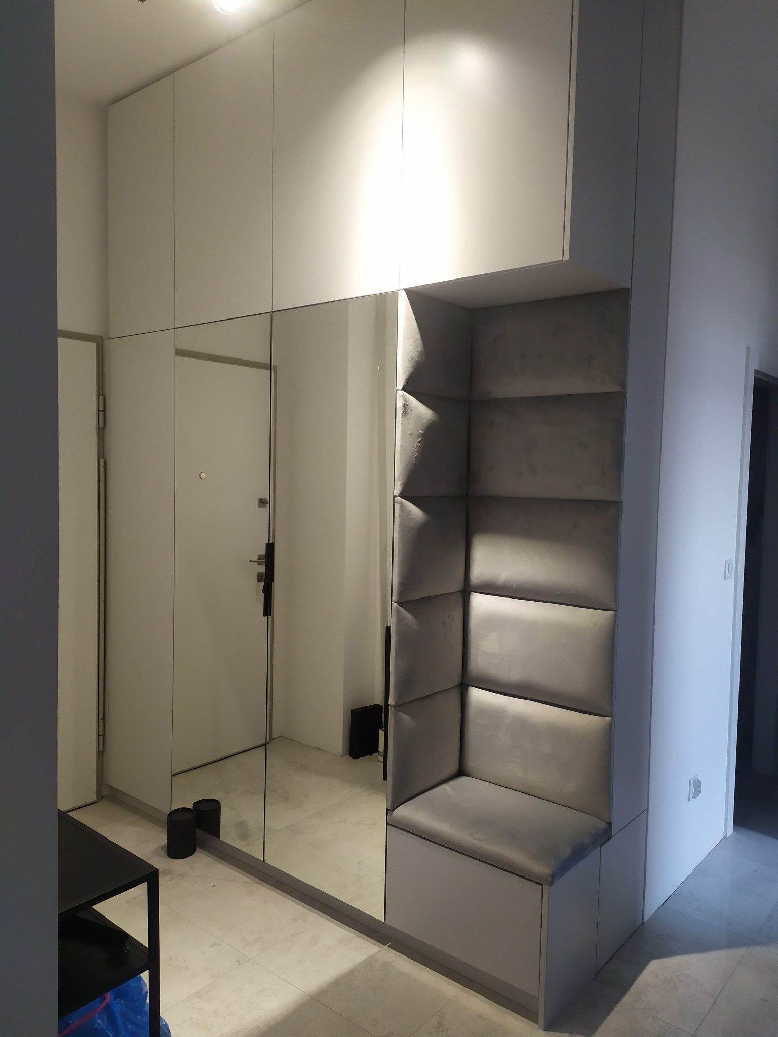 Wnętrze przedpokoju z szafą w zabudowie, lustrem i tapicerowanym siedziskiem w rogu, odbicie drzwi wejściowych w lustrze.