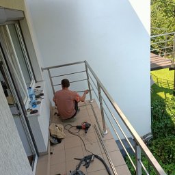 Loft d. S. - Montaż balustrady ze stali nierdzewnej na balkonie, widoczny pracownik w trakcie pracy, narzędzia i elementy konstrukcyjne na podłodze, w tle zielona roślinność i schody zewnętrzne.