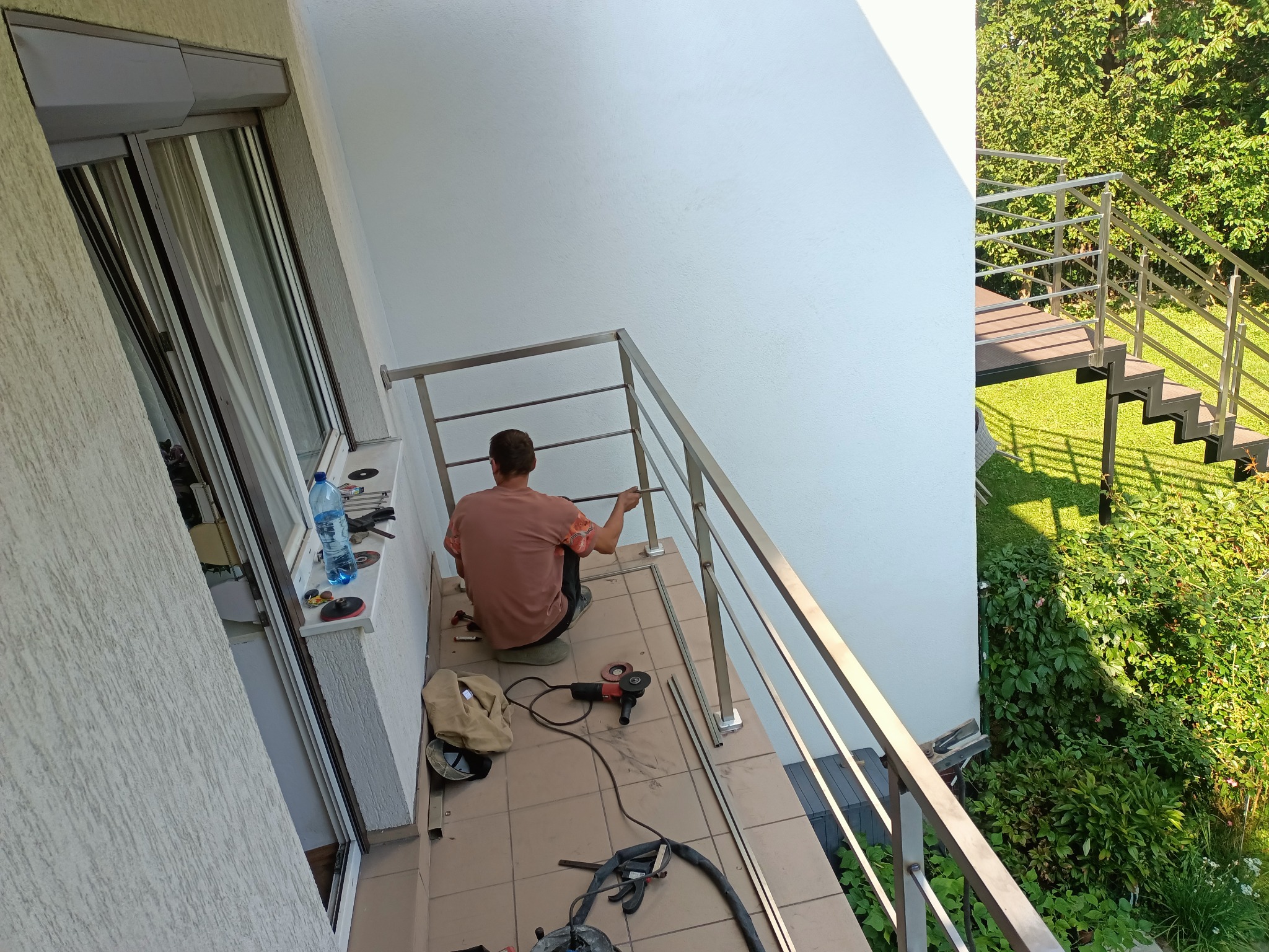 Montaż balustrady ze stali nierdzewnej na balkonie, widoczny pracownik w trakcie pracy, narzędzia i elementy konstrukcyjne na podłodze, w tle zielona roślinność i schody zewnętrzne.