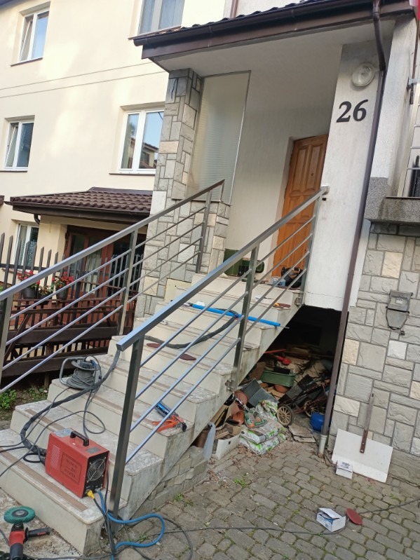 Montaż balustrady ze stali nierdzewnej przy schodach wejściowych do domu z numerem 26, widoczne narzędzia i materiały budowlane.