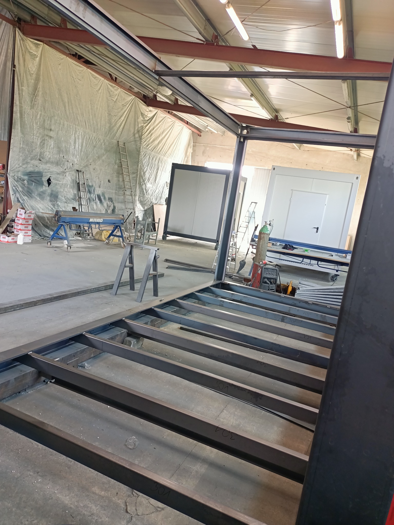 Wnętrze hali produkcyjnej z widocznym szkieletem konstrukcji stalowej pod budynek modułowy, z narzędziami i materiałami w tle.