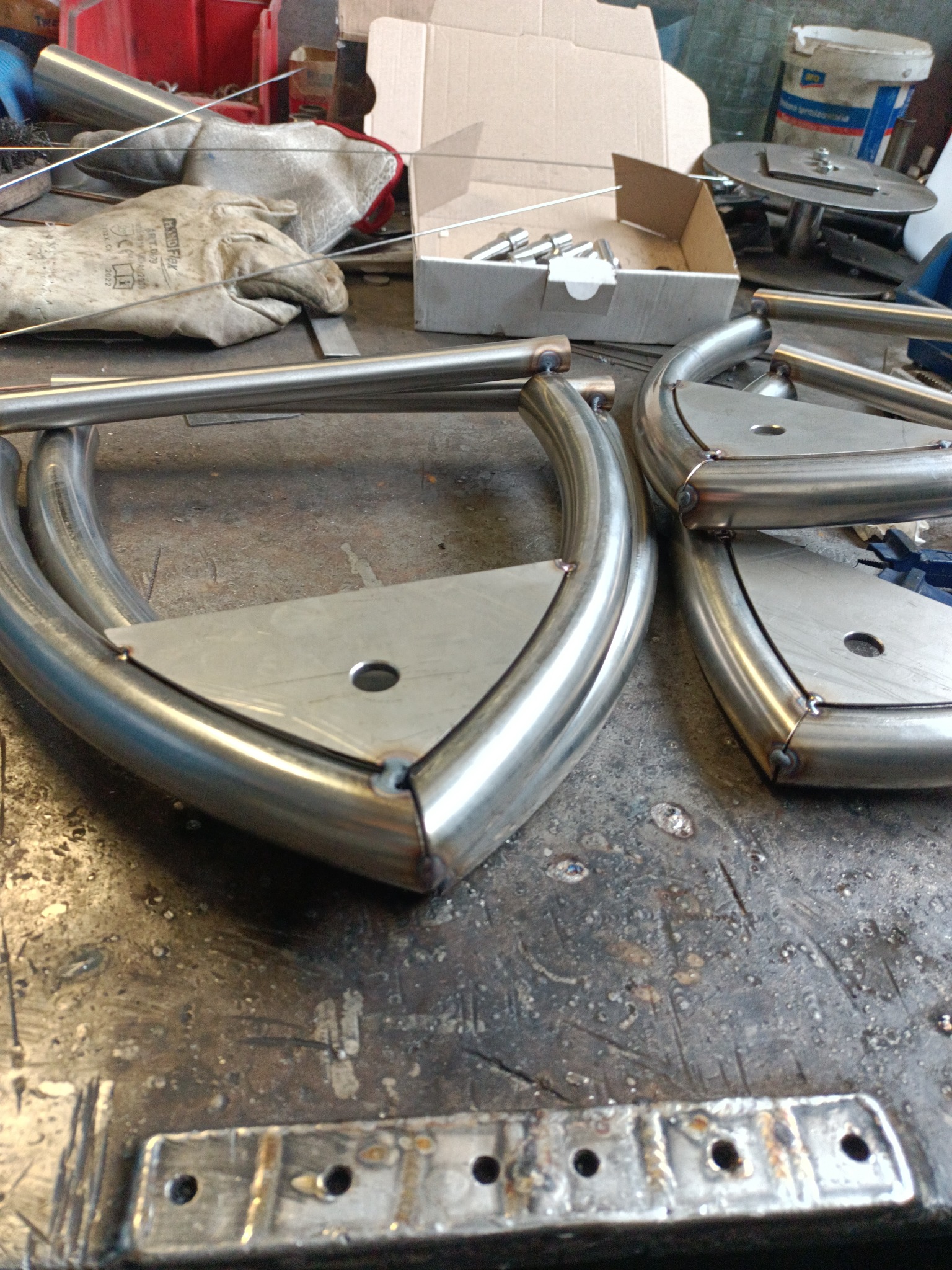 Dwa półokrągłe elementy konstrukcyjne ze stali nierdzewnej, połączone spawanymi, trójkątnymi płytkami z otworem, leżące na brudnym stole warsztatowym z narzędziami i akcesoriami w tle.