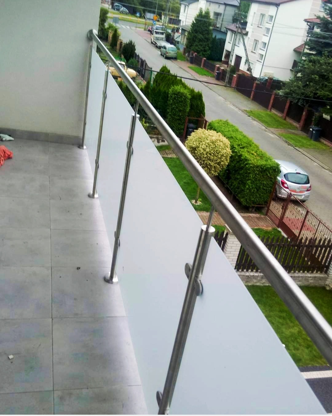 Nowoczesna balustrada balkonowa ze szkła mlecznego i stali nierdzewnej, widok z góry na ulicę z zielenią.