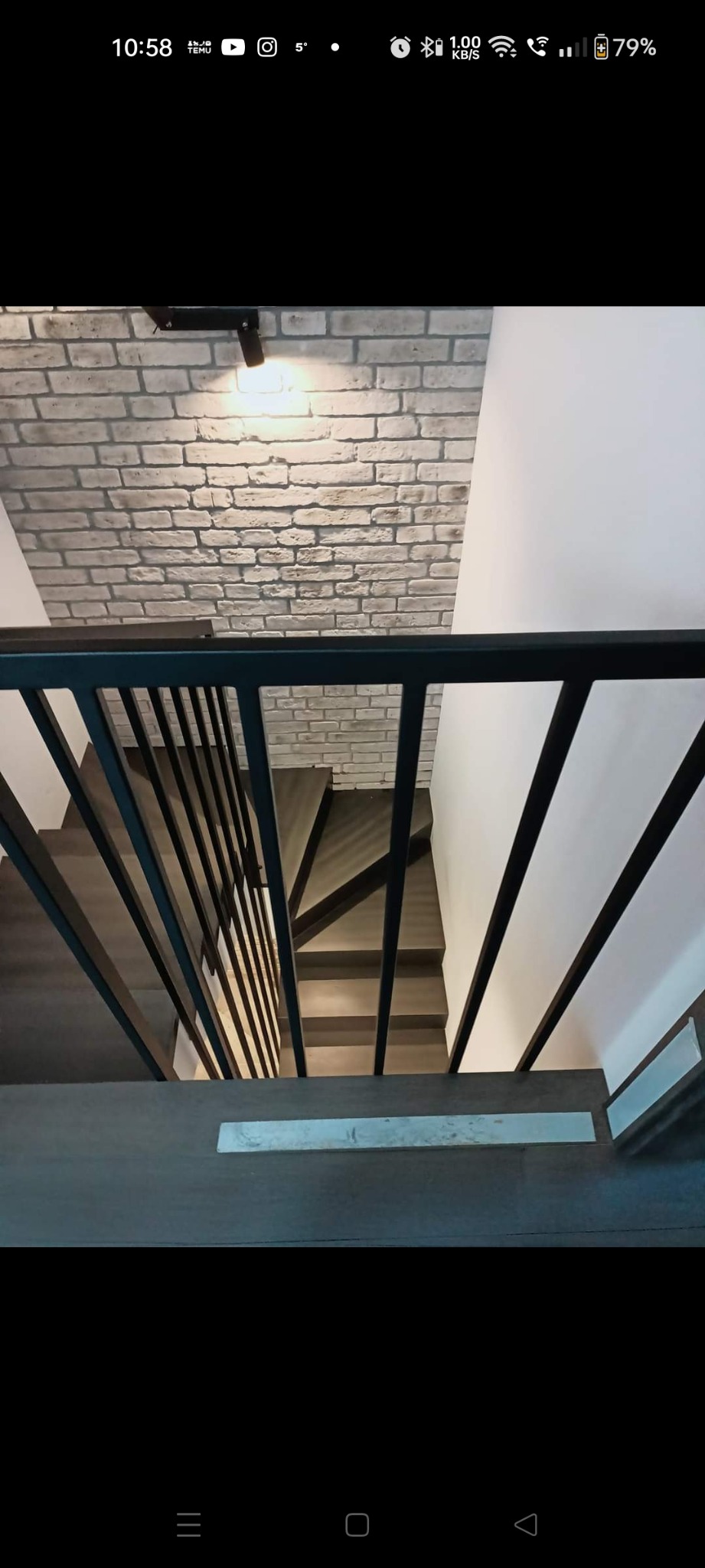 Widok z góry na schody z ciemnego drewna z czarną metalową balustradą, ściana wykończona cegłą w tle.