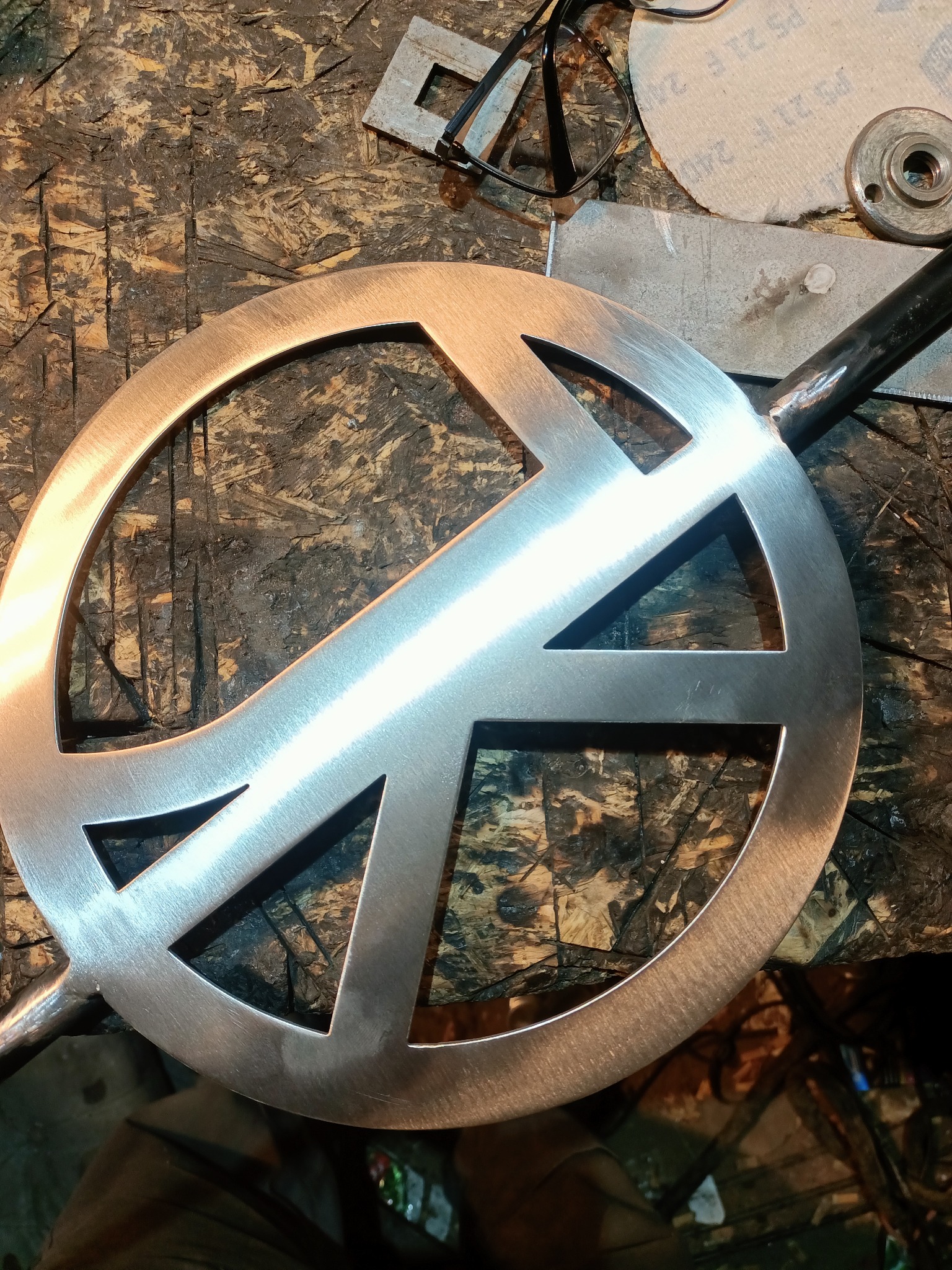 Metalowy element w kształcie znaku pokoju, z przyspawanym prętem, leży na warsztatowym stole. Widoczne ślady obróbki i narzędzia.
