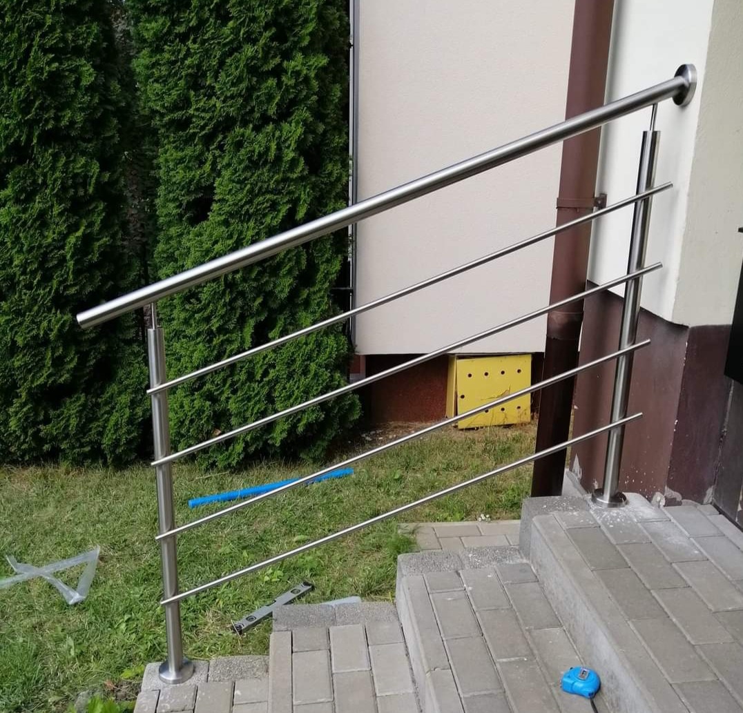 Zewnętrzna balustrada schodowa ze stali nierdzewnej z czterema poziomymi prętami, zamontowana przy wejściu do budynku z zielonymi tujami w tle.