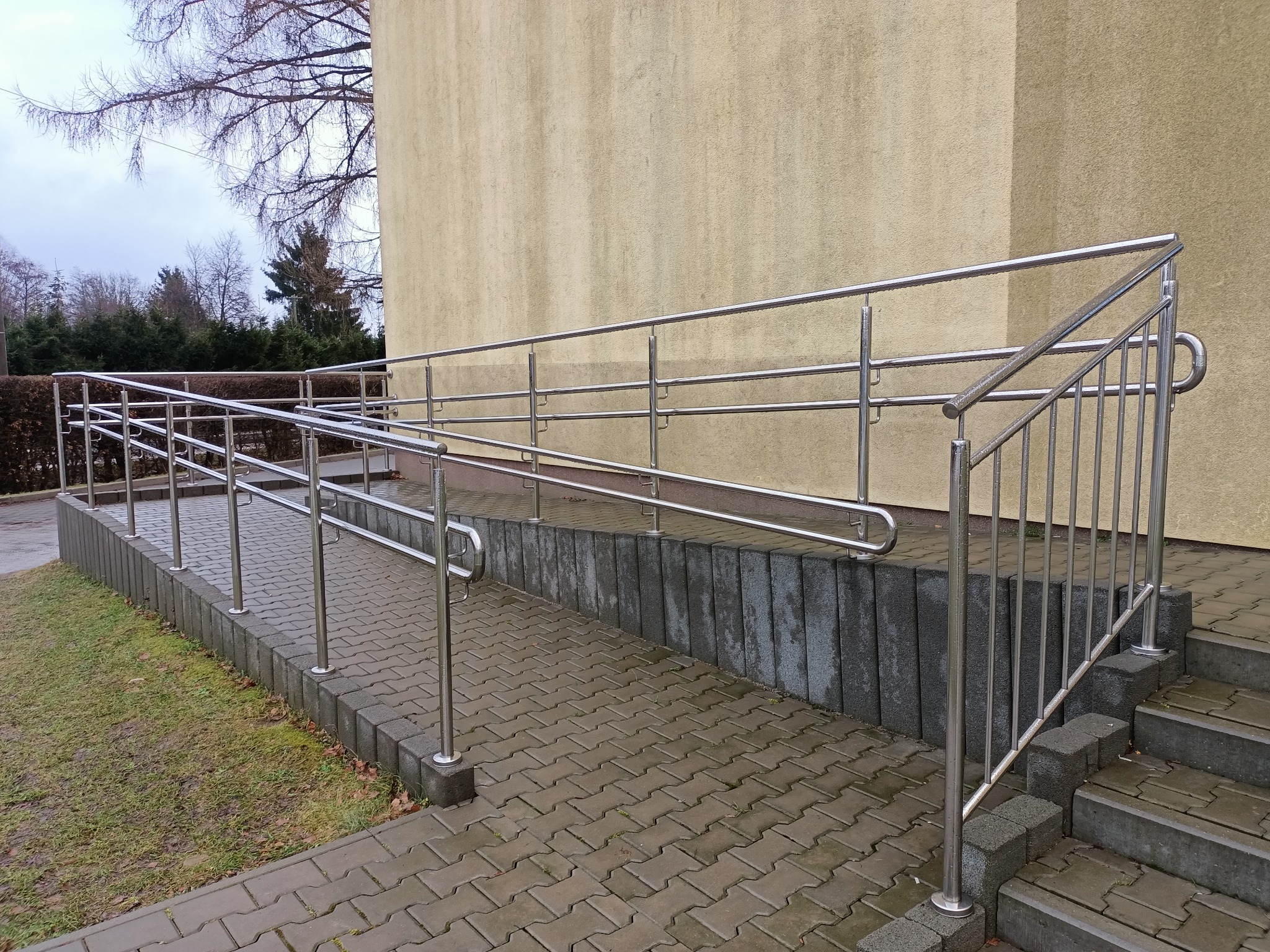 Zewnętrzna, metalowa poręcz ze stali nierdzewnej, zamontowana przy schodach i podjeździe dla wózków inwalidzkich, na tle beżowej ściany budynku i kostki brukowej.