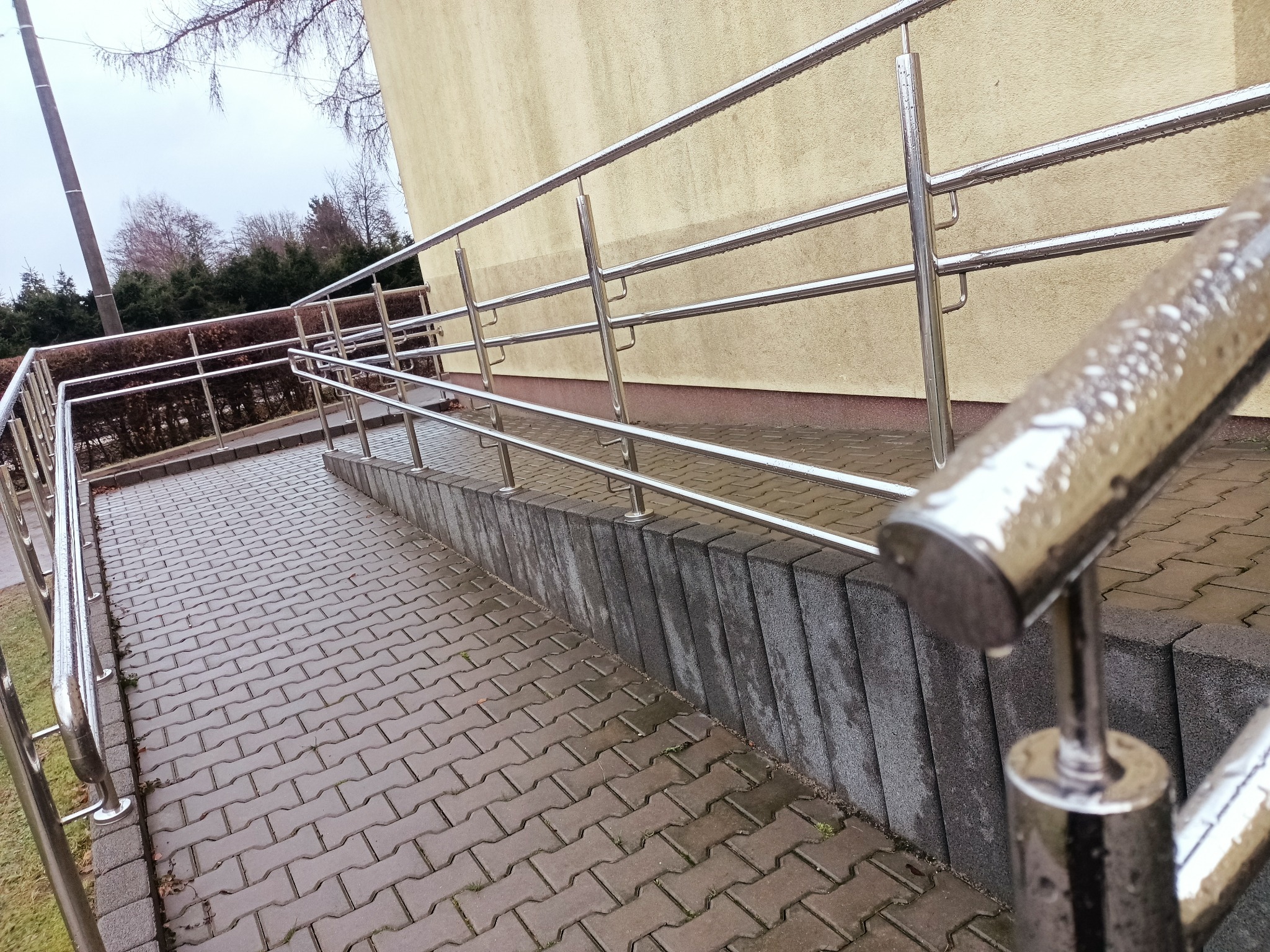 Zewnętrzna rampa wejściowa z nierdzewną balustradą, widoczne krople wody po deszczu, kostka brukowa i fragment budynku w tle.