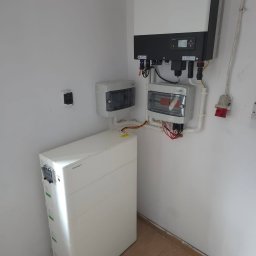 SUNUPS DANIEL ŻESŁAWSKI - Biały magazyn energii Growatt, inwerter i rozdzielnice na ścianie w pomieszczeniu. Widoczne kable i instalacja elektryczna. Ujęcie z dołu, z perspektywy podłogi.