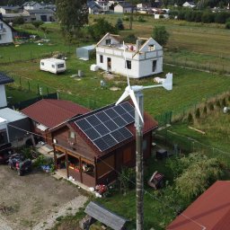SUNUPS DANIEL ŻESŁAWSKI - Dom z panelami słonecznymi na dachu i turbiną wiatrową na posesji. Widok z lotu ptaka na budynek z brązowego drewna i czerwoną dachówką w otoczeniu zieleni.