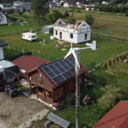 Dom z panelami słonecznymi na dachu i turbiną wiatrową na posesji. Widok z lotu ptaka na budynek z brązowego drewna i czerwoną dachówką w otoczeniu zieleni.
