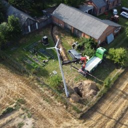 SUNUPS DANIEL ŻESŁAWSKI - Montaż turbiny wiatrowej na polu, widok z drona. Ekipa montażowa, wykopany dół, dźwig i ciężarówka. W tle budynki gospodarcze i ogród warzywny.