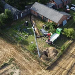 Montaż turbiny wiatrowej na polu, widok z drona. Ekipa montażowa, wykopany dół, dźwig i ciężarówka. W tle budynki gospodarcze i ogród warzywny.