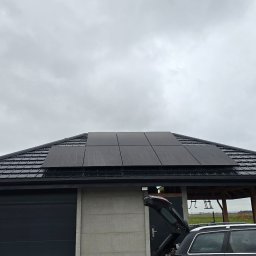 SUNUPS DANIEL ŻESŁAWSKI - Czarny dach z panelami słonecznymi w pochmurny dzień. Widoczny garaż z szarą elewacją, fragment samochodu z otwartym bagażnikiem i zadaszona konstrukcja w tle.