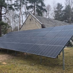 SUNUPS DANIEL ŻESŁAWSKI - Panel fotowoltaiczny na metalowej konstrukcji, ustawiony na trawniku przed budynkiem gospodarczym z szarej cegły. W tle drzewa i fragment betonowego ogrodzenia.