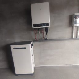 SUNUPS DANIEL ŻESŁAWSKI - Biały magazyn energii z podłączonym inwerterem na szarej ścianie. Widoczne kable, skrzynka elektryczna i fragment niebieskiej rury. Minimalistyczny design.