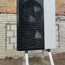 Zewnętrzna pompa ciepła Hewalex z dwoma wentylatorami, zamontowana na białym stelażu przy ceglanym budynku.