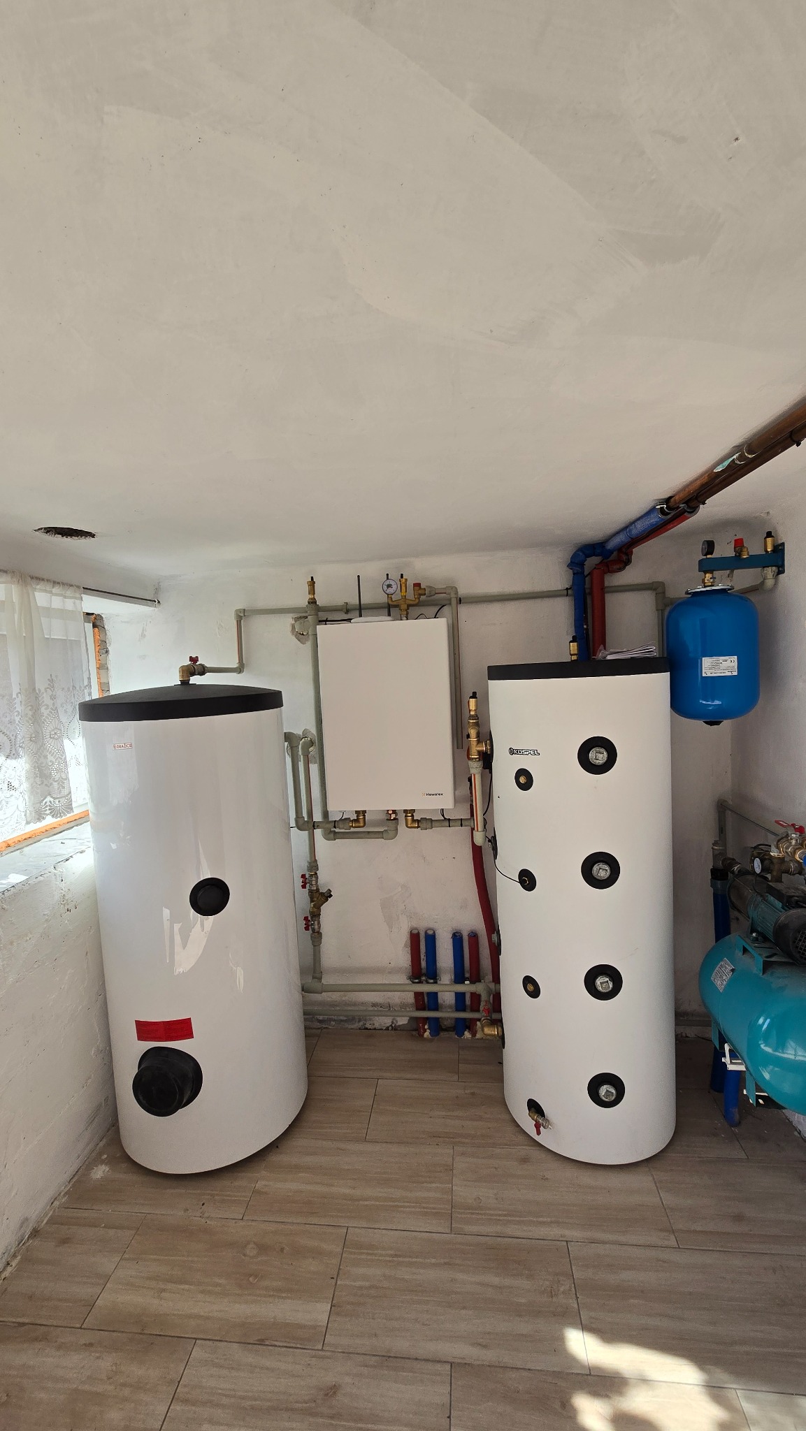 Dwa białe zbiorniki akumulacyjne ciepła, instalacja grzewcza z rurami, manometrem i zaworami w pomieszczeniu z beżową podłogą i oknem z zasłoną.