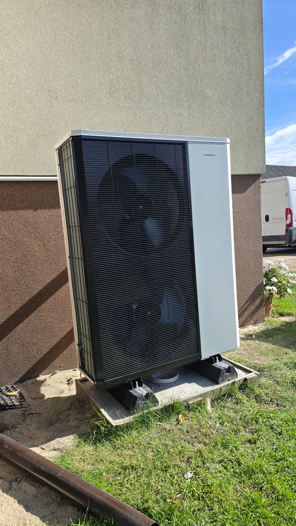 Pompa ciepła Hewalex zamontowana przy ścianie budynku, widoczna instalacja na betonowej podstawie, trawa i fragment rynny. Biały bus w tle.