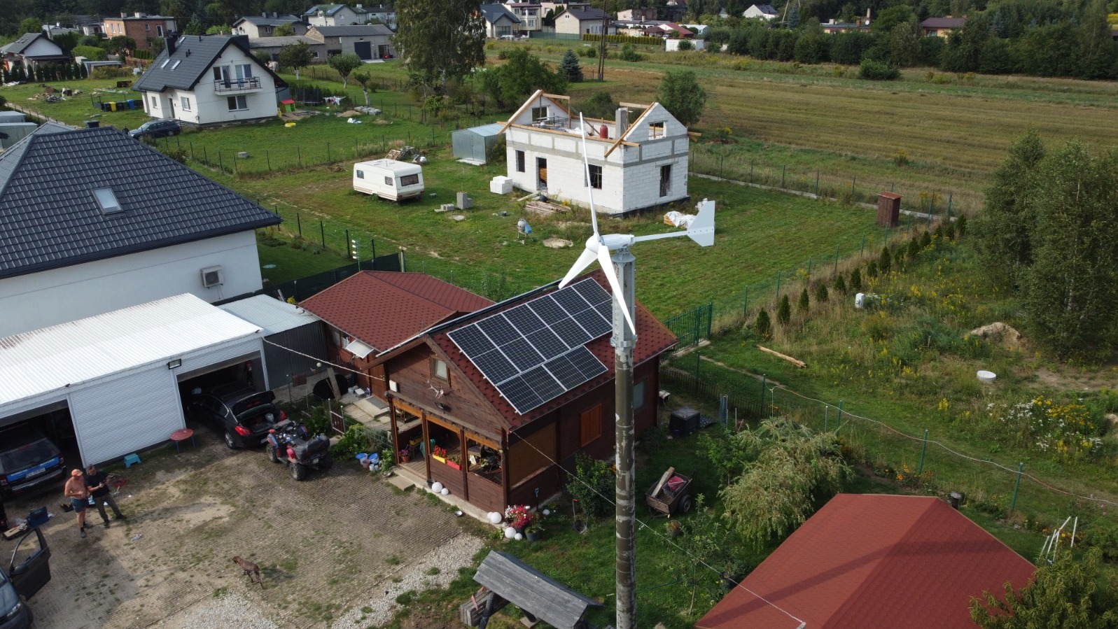 Dom z panelami słonecznymi na dachu i turbiną wiatrową na posesji. Widok z lotu ptaka na budynek z brązowego drewna i czerwoną dachówką w otoczeniu zieleni.