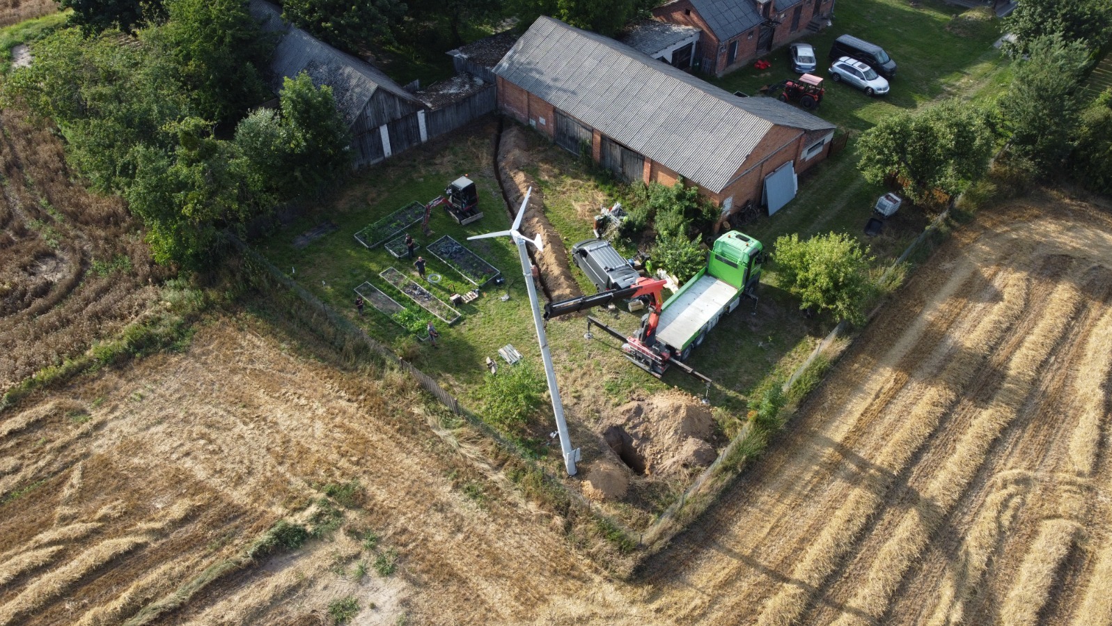 Montaż turbiny wiatrowej na polu, widok z drona. Ekipa montażowa, wykopany dół, dźwig i ciężarówka. W tle budynki gospodarcze i ogród warzywny.
