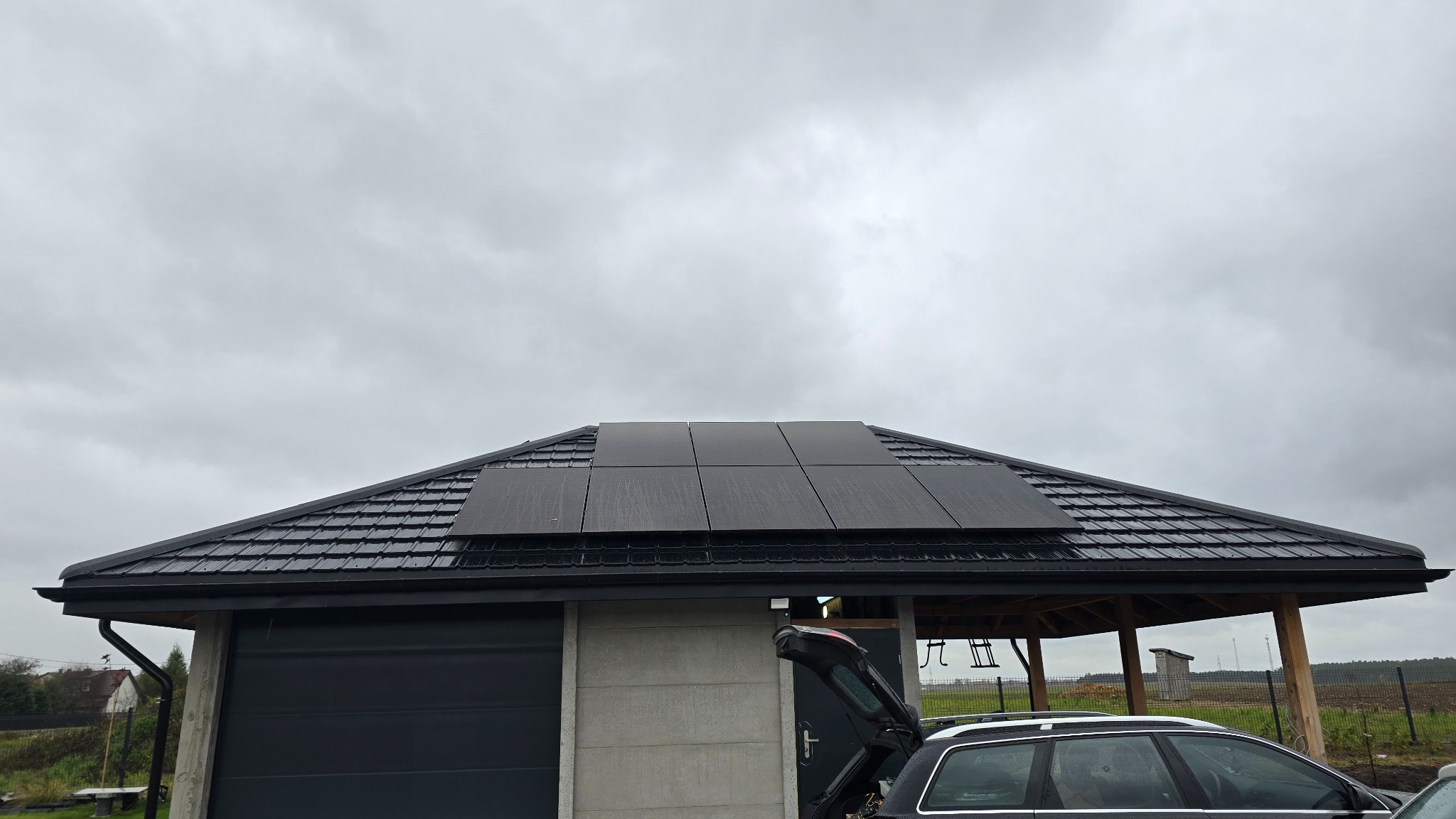 Czarny dach z panelami słonecznymi w pochmurny dzień. Widoczny garaż z szarą elewacją, fragment samochodu z otwartym bagażnikiem i zadaszona konstrukcja w tle.