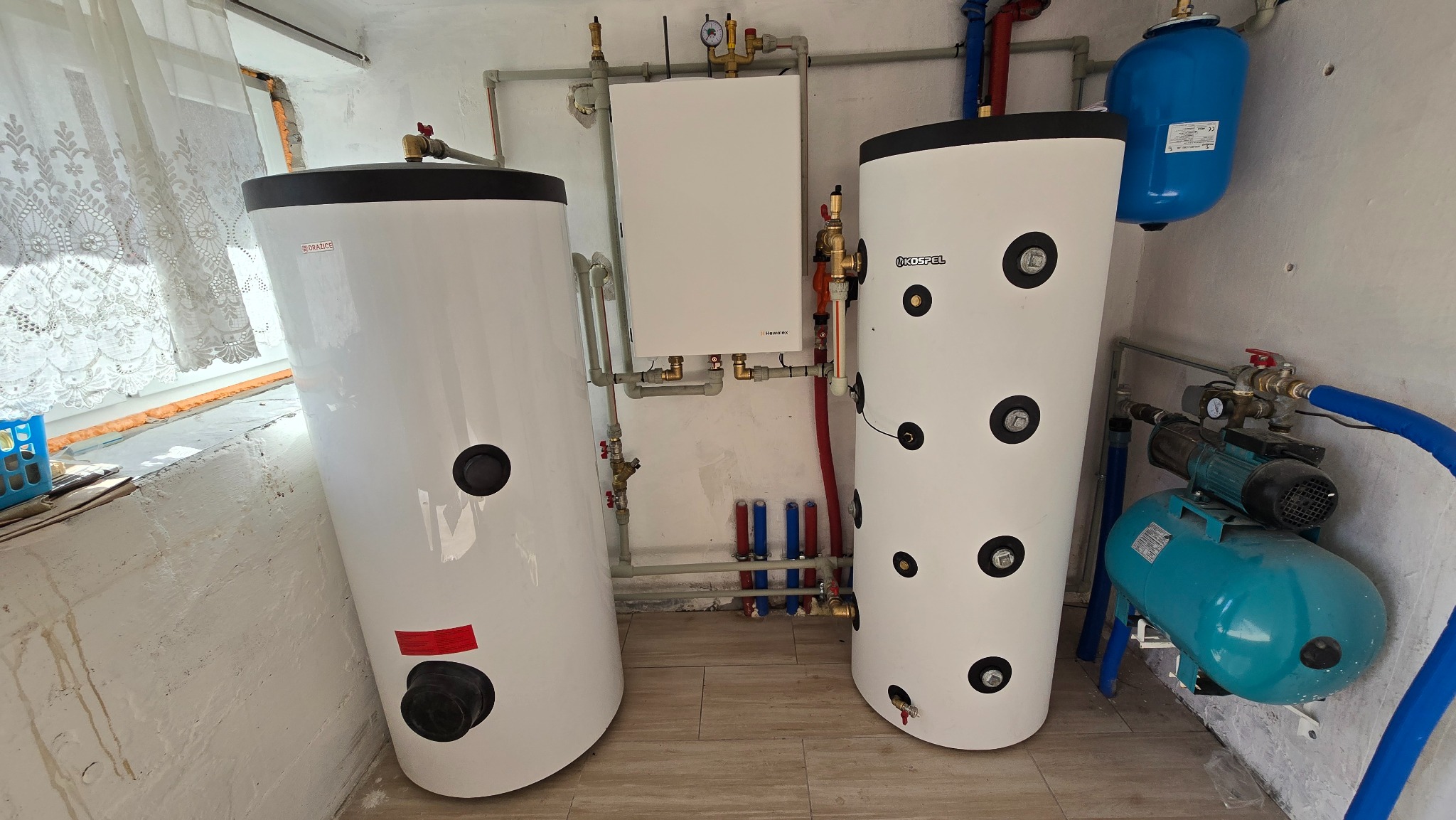 Instalacja dwóch białych zbiorników ciepłej wody użytkowej (CWU) marek Drazice i Kospel, z piecem grzewczym Hewalex i niebieskim zbiornikiem hydroforowym w kotłowni.