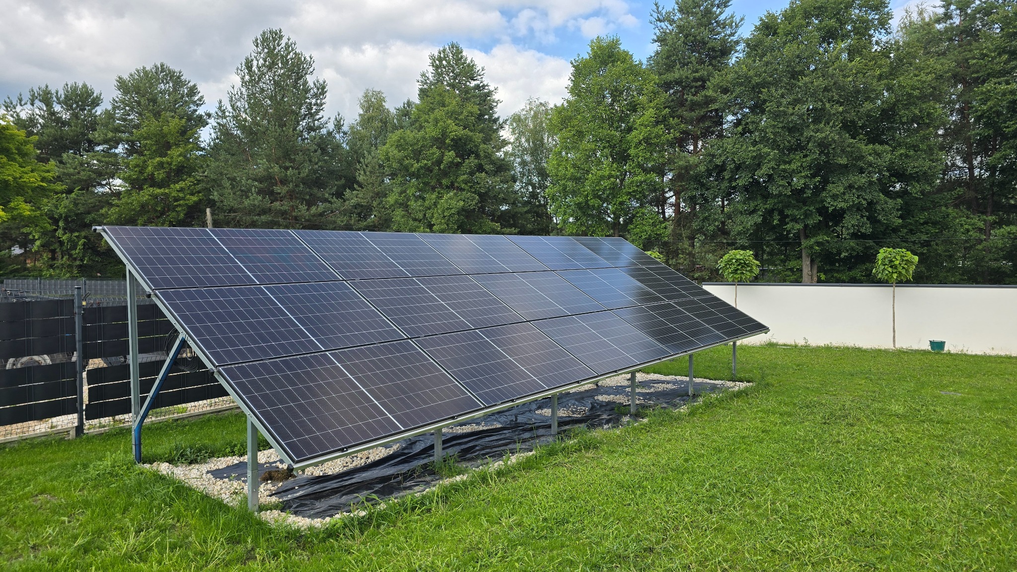 Panel słoneczny na metalowej konstrukcji, otoczony zieloną trawą i drzewami. Widoczne niebo z chmurami. Instalacja na podłożu z kamieni i czarnej folii.
