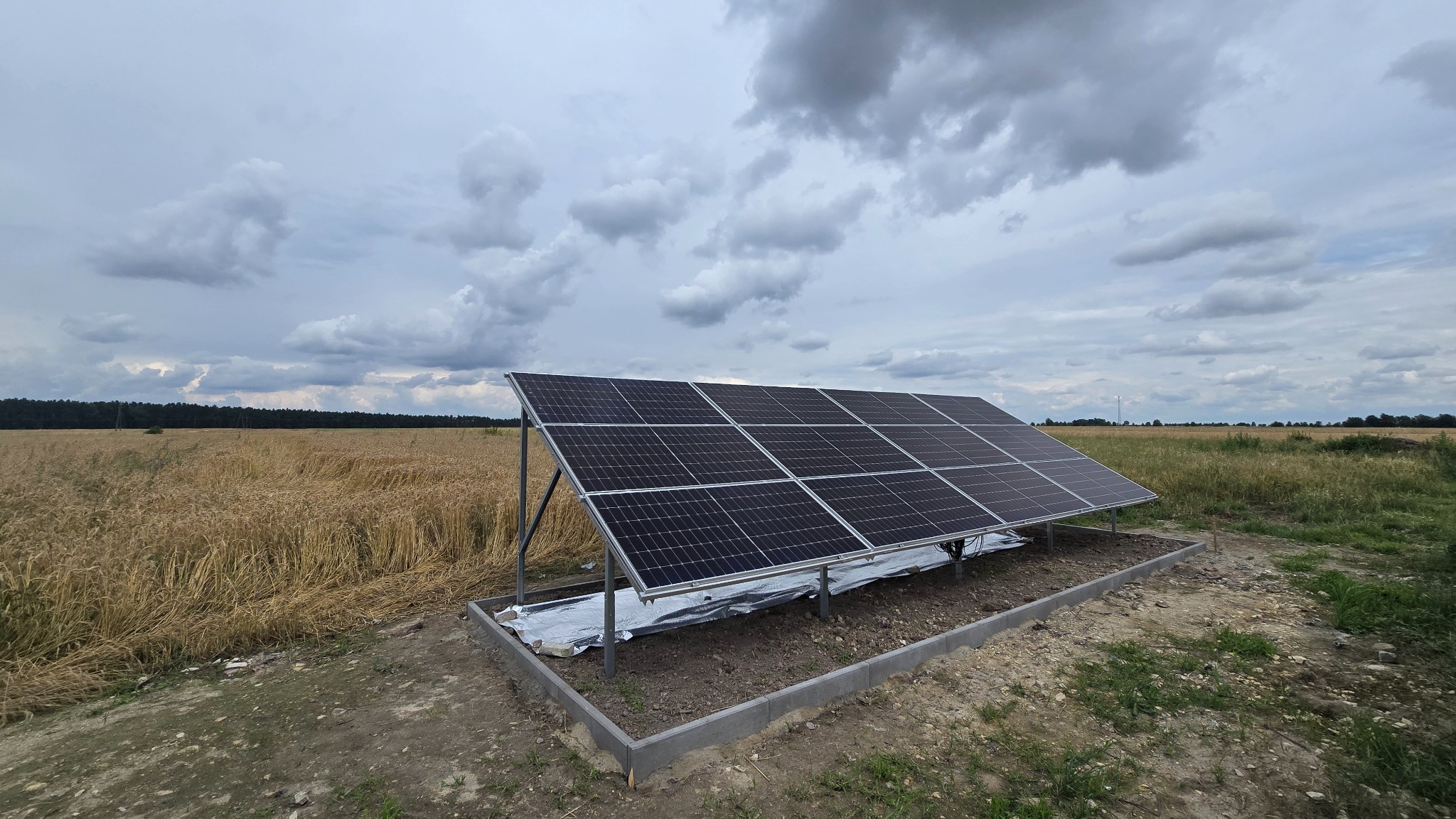 Panel słoneczny na metalowej konstrukcji, otoczony betonową obwódką, na tle pola i pochmurnego nieba. Instalacja w toku, zabezpieczona folią.