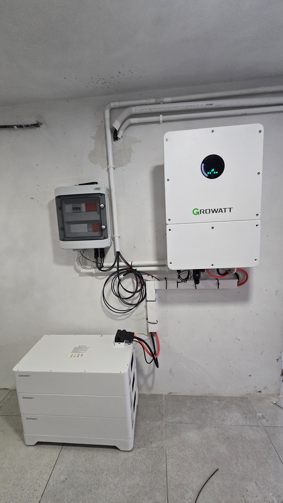 Biały inwerter Growatt z podłączonymi kablami, skrzynka z bezpiecznikami i modułowa bateria na szarej podłodze. Instalacja w pomieszczeniu o surowym wykończeniu.