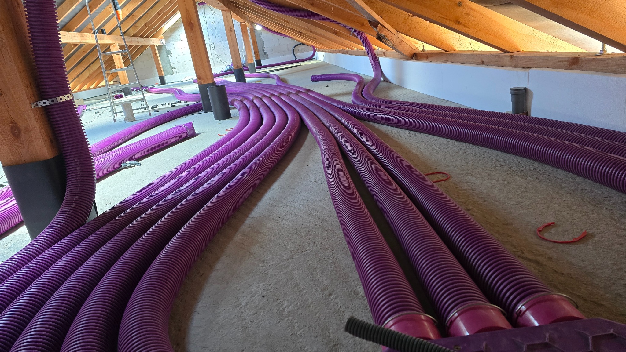 Fioletowe rury rekuperacji ułożone na poddaszu, widoczne drewniane elementy konstrukcji dachu i drabina. Instalacja wentylacyjna w trakcie montażu.