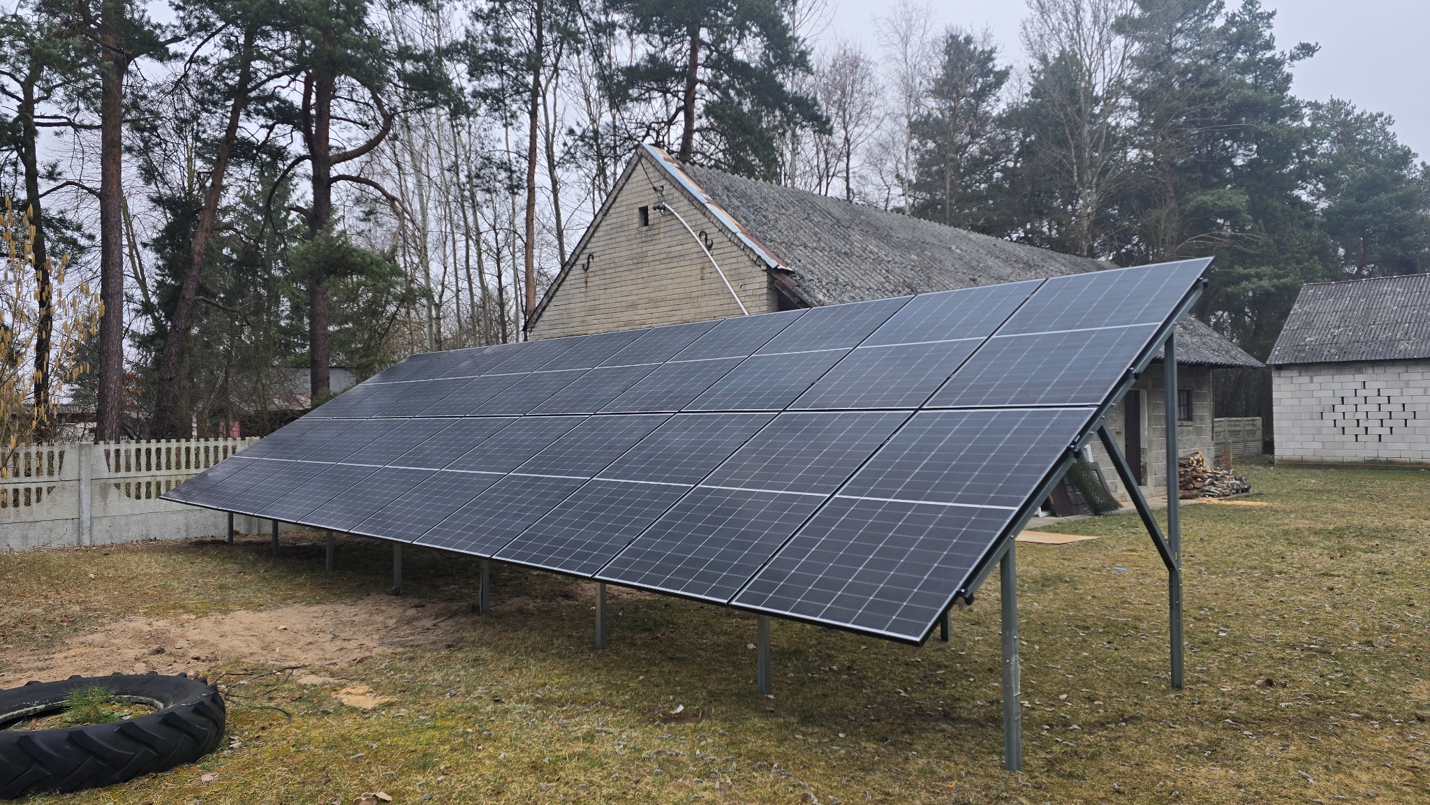 Panel fotowoltaiczny na metalowej konstrukcji, ustawiony na trawniku przed budynkiem gospodarczym z szarej cegły. W tle drzewa i fragment betonowego ogrodzenia.