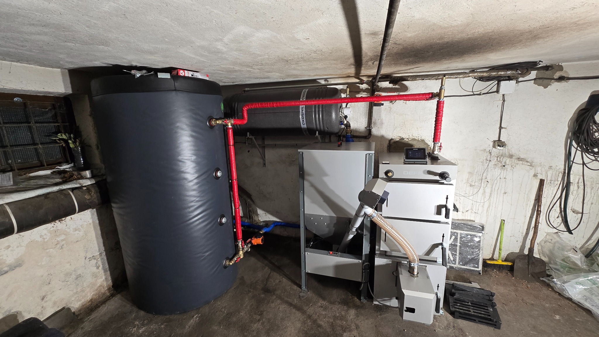 Zestaw kotła na pellet z zasobnikiem i zbiornikiem buforowym w kotłowni. Widoczne rury w izolacji termicznej. Instalacja grzewcza w piwnicy.