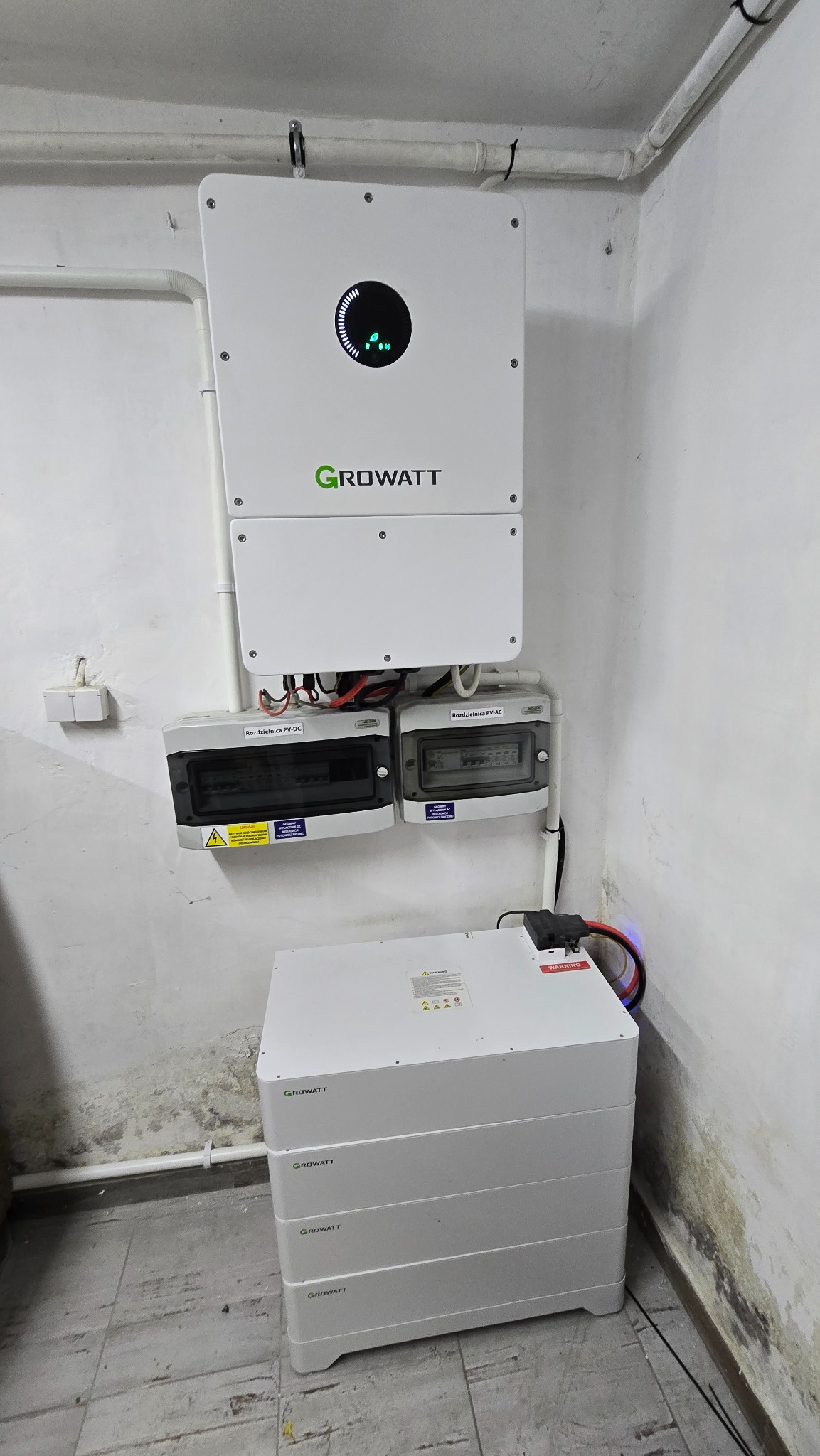 Biały inwerter Growatt z rozdzielnicami PV-DC i PV-AC oraz modułowy magazyn energii w pomieszczeniu. Widoczne okablowanie i elementy instalacji.