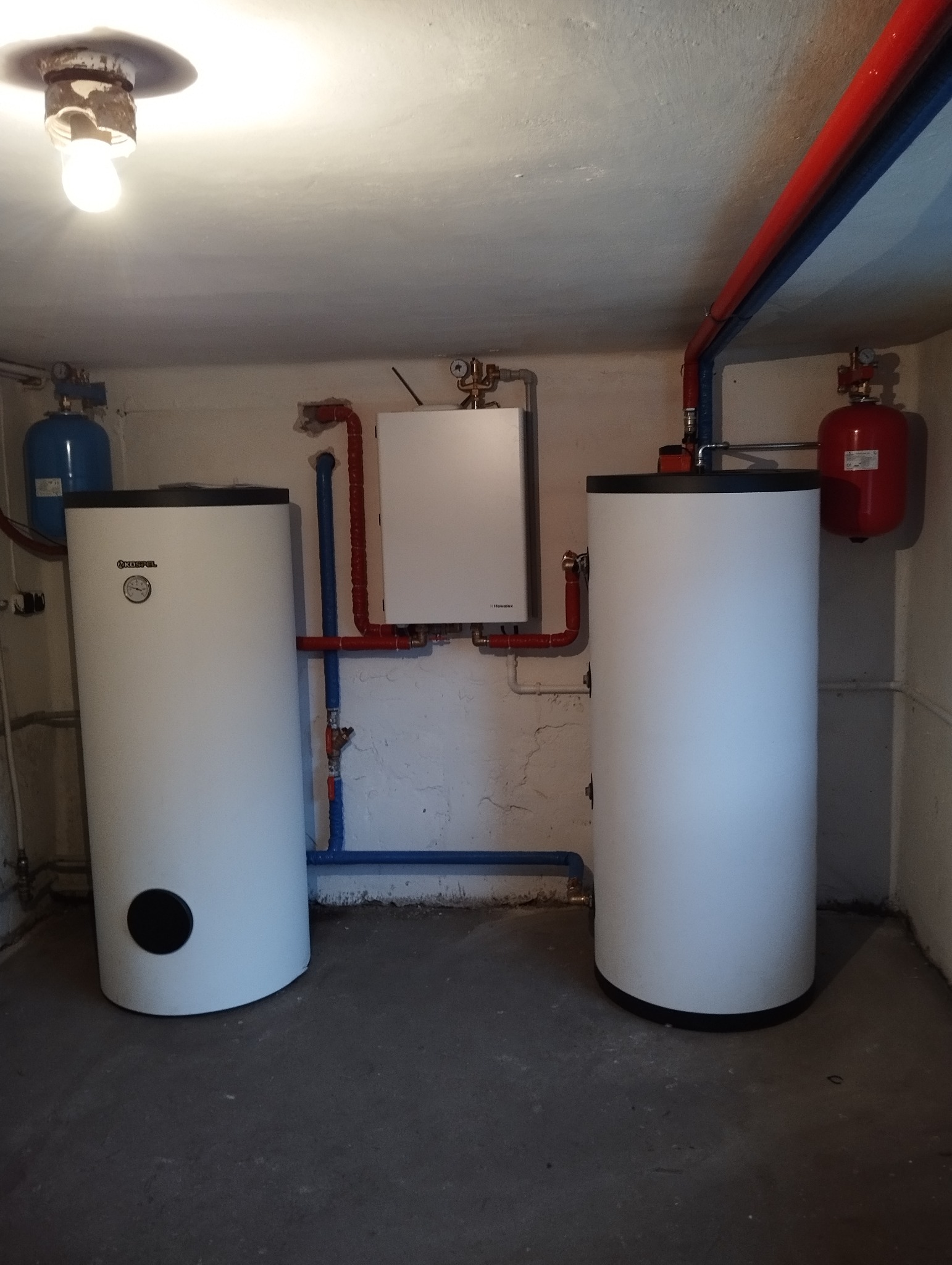 Dwa białe zbiorniki akumulacyjne Kospel w piwnicy, połączone rurami w kolorach czerwonym i niebieskim, z małym białym kotłem wiszącym na ścianie i czerwonym zbiornikiem wyrównawczym.